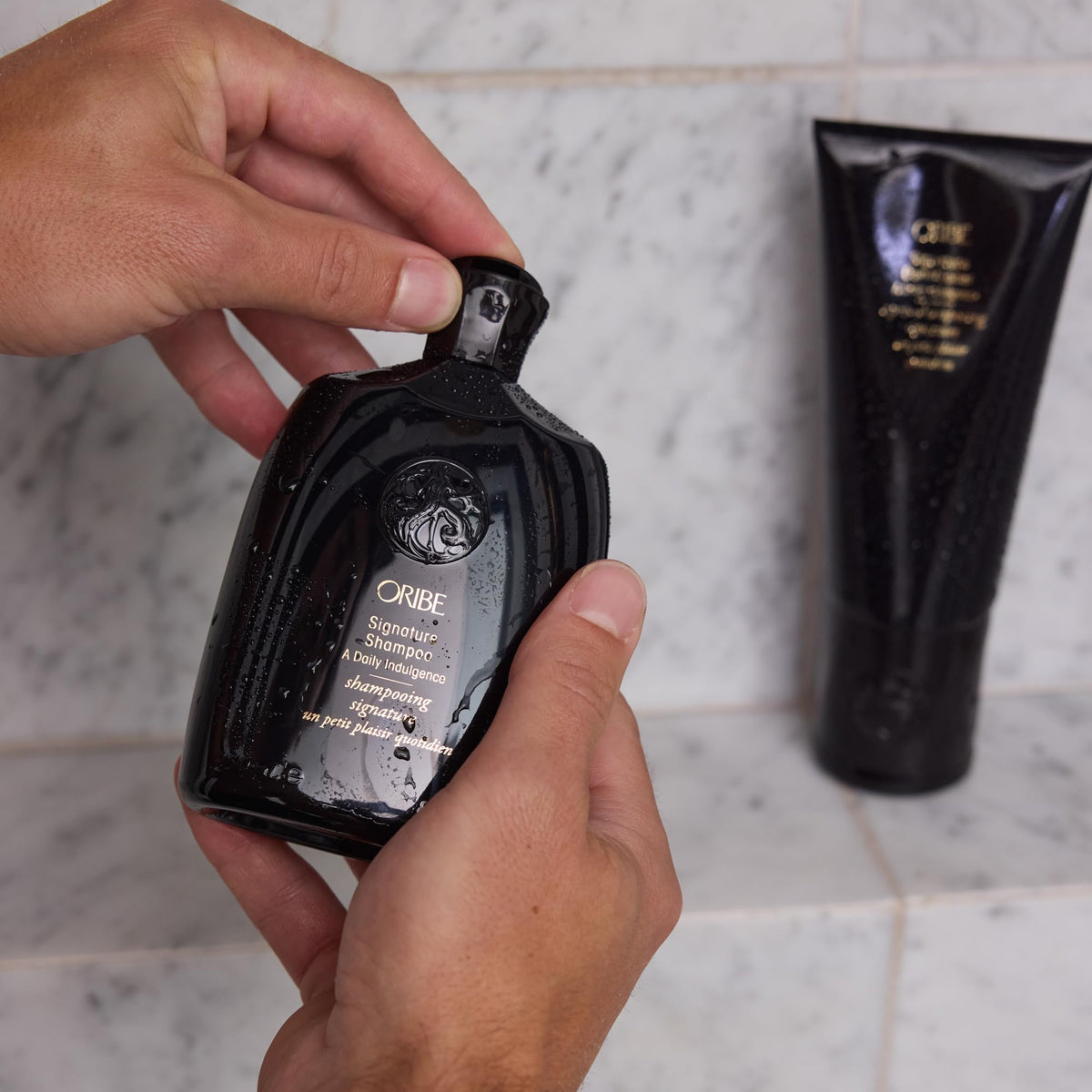 ORIBE bottle in hand under shower illustrating rinsable, weightless cleansing benefit
